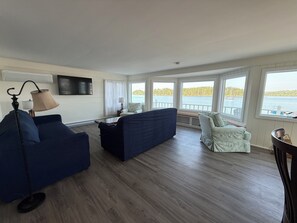 A living room with a sofa, armchair, and a table. There are windows with a view of the water and trees. - Heron's Landing (Harpswell)