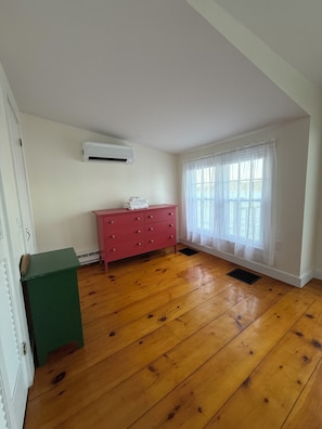 Una habitación con piso de madera, un aparador rosa, un equipo de aire acondicionado blanco y una ventana con cortinas translúcidas.