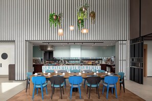 A modern dining area with a long table, blue chairs, hanging plants, and a kitchen in the background. - Courtyard By Marriott Dakar Diamniadio (Diamniadio)