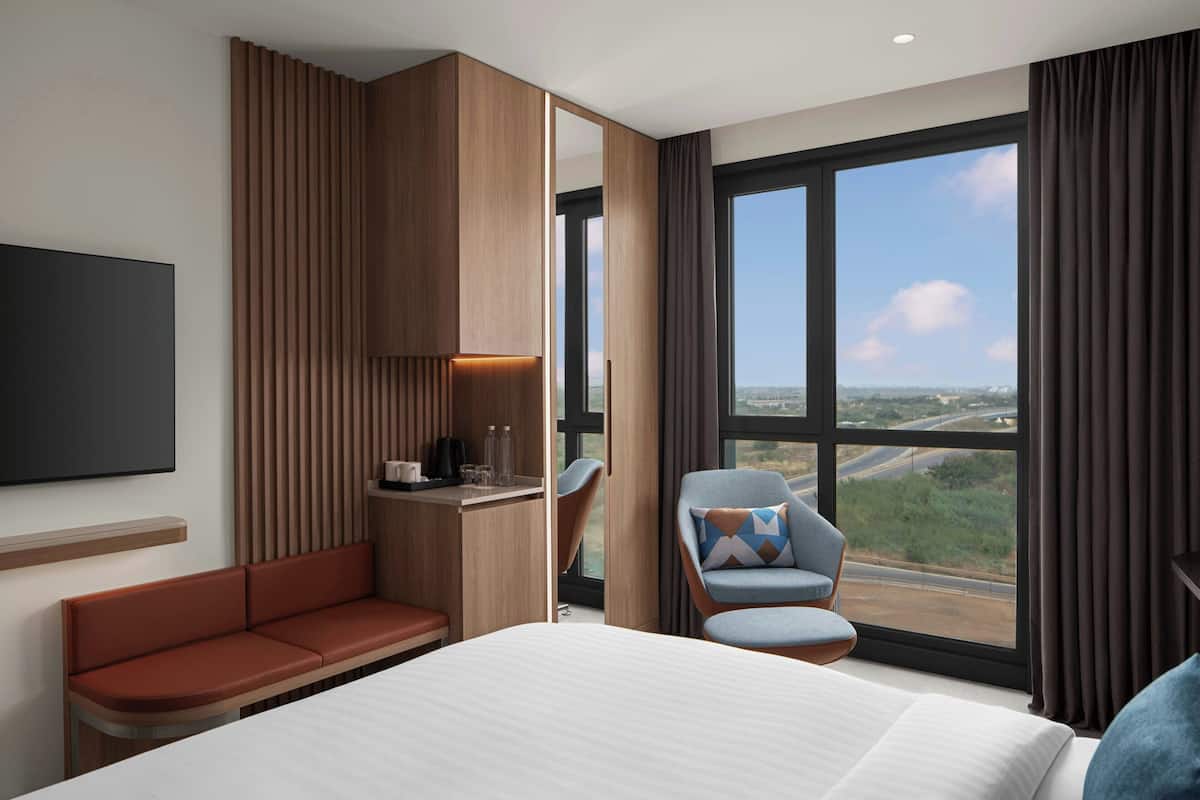 A modern hotel room with a large bed, a seating area, a desk, and a view of the city.