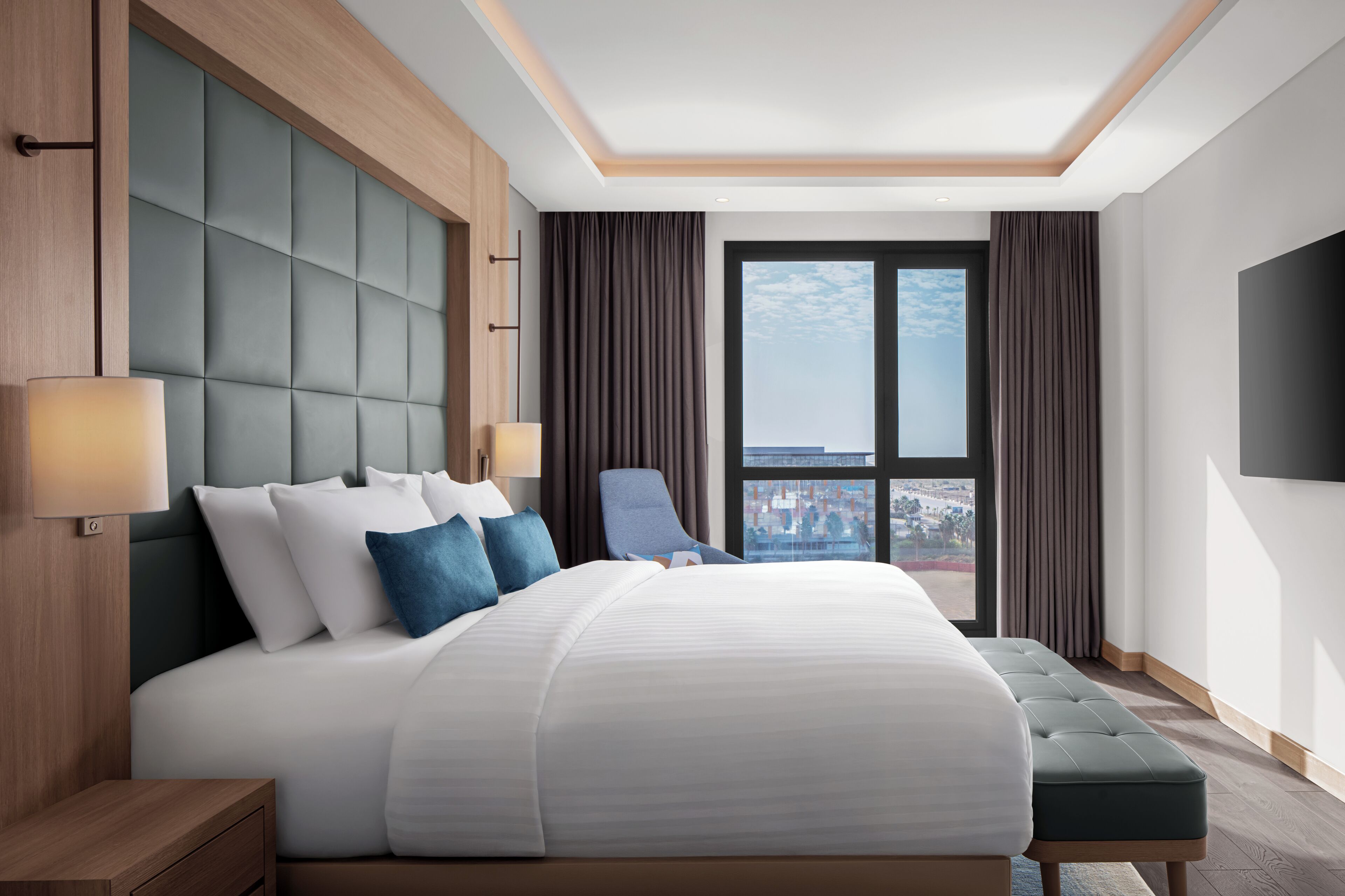 A modern hotel room with a large bed, a view of the city, and a flat-screen TV on the wall.