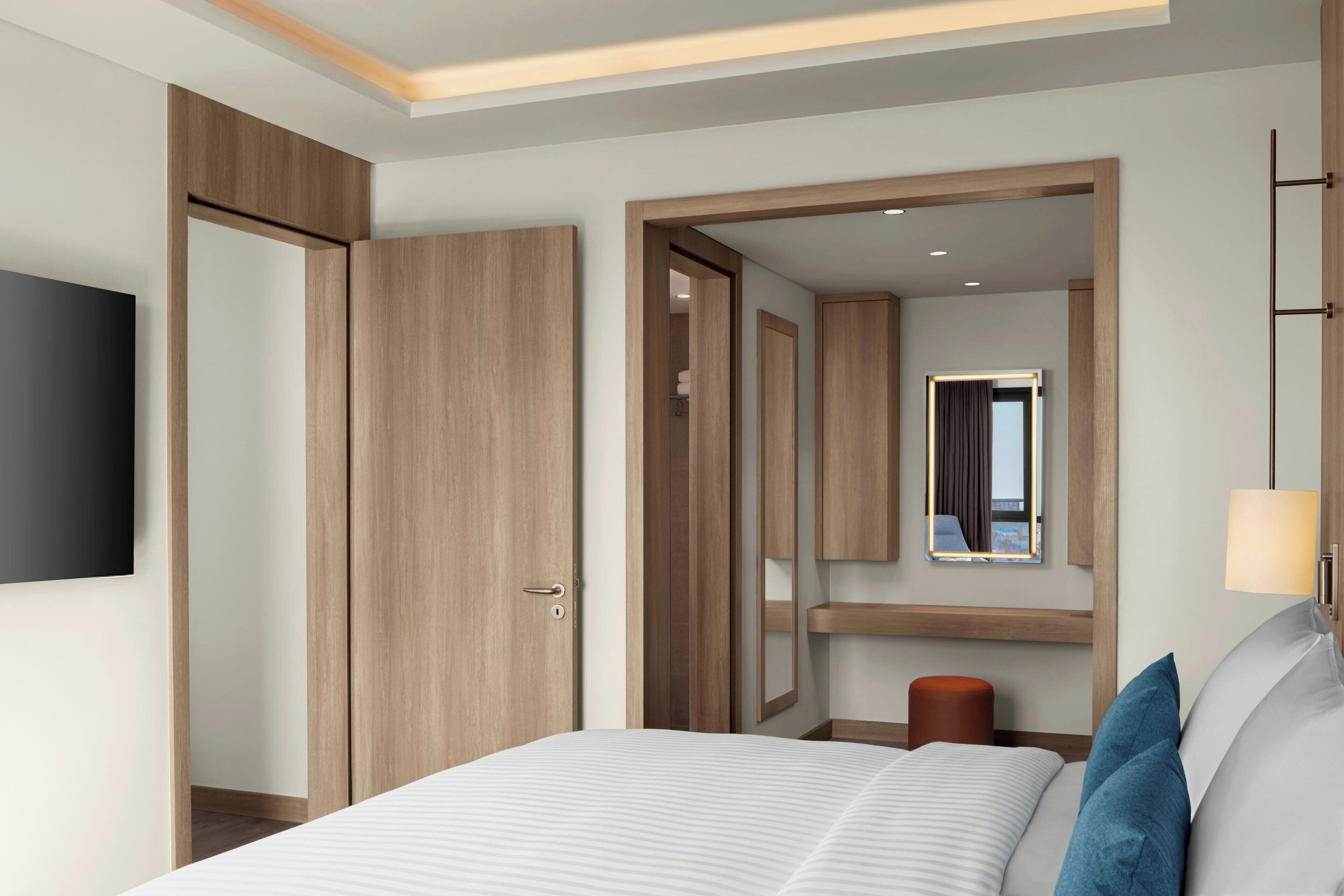 A modern hotel room with a large bed, a flat-screen TV, wooden wardrobes, a mirror, and a bedside lamp.