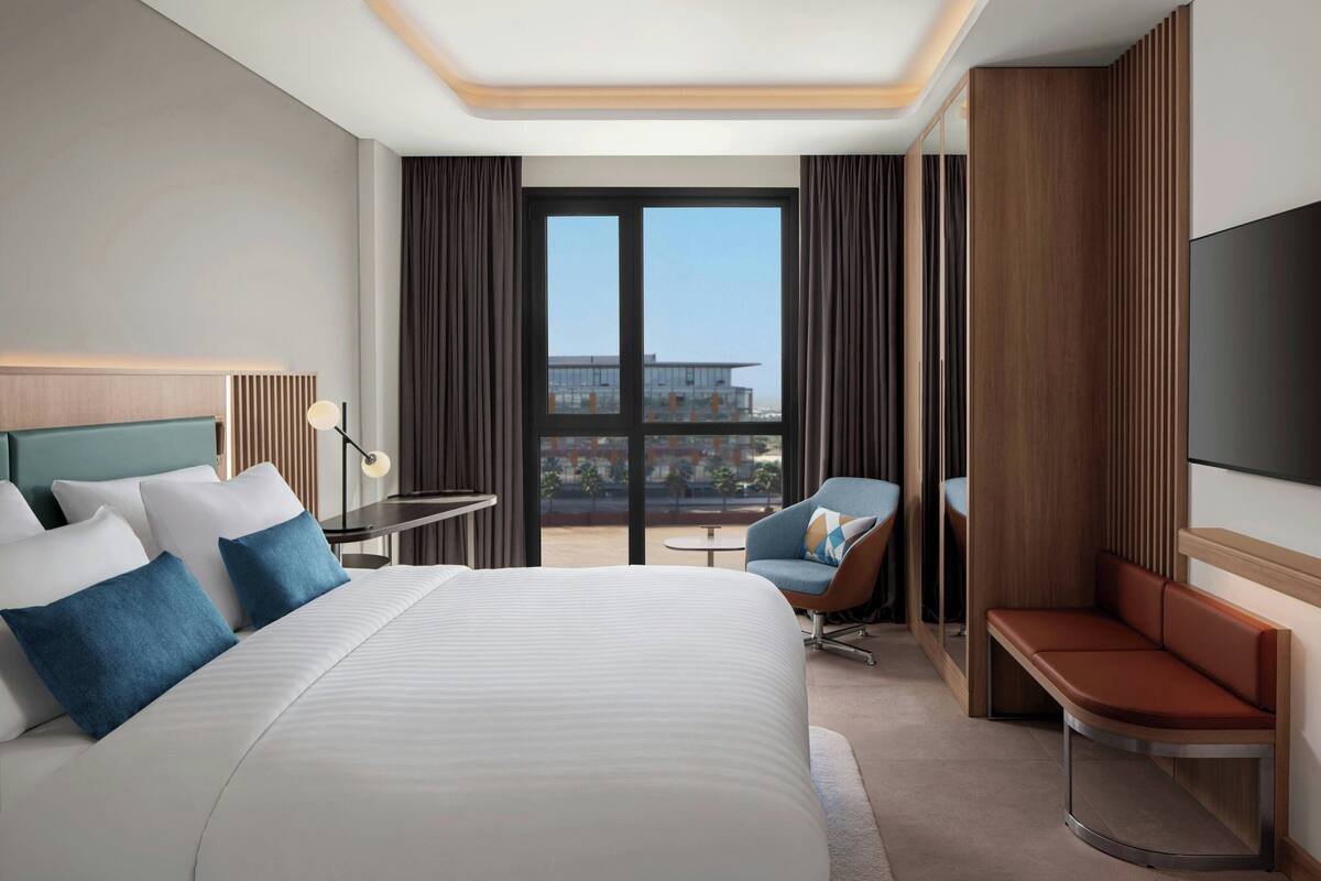 A hotel room with a large bed, a desk with a lamp, a chair, a sofa, and a TV on the wall.