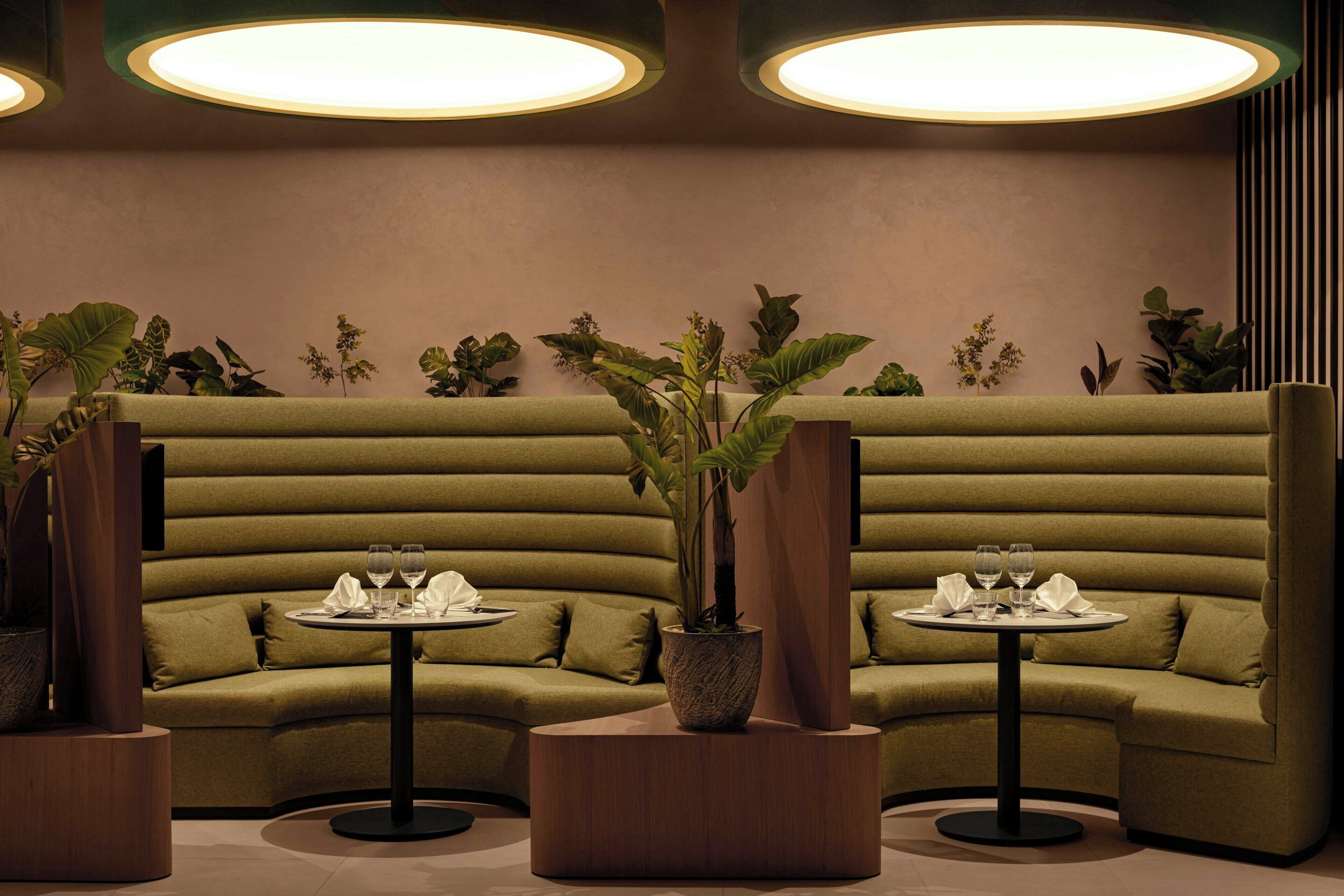 A modern lounge area with green upholstered seating, two round tables with wine glasses, and potted plants.