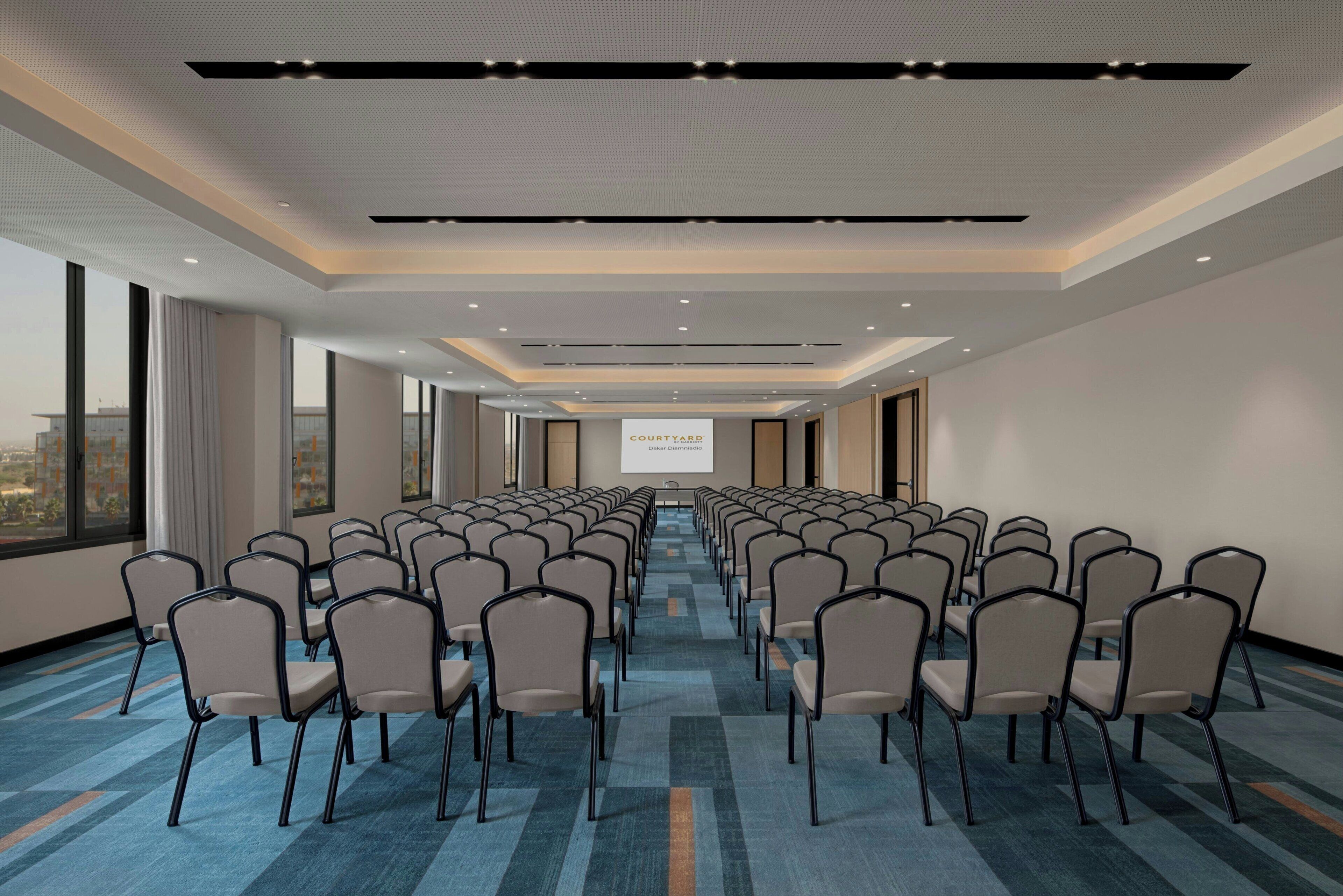 Un salón de conferencias con filas de sillas, una pantalla de proyector y un amplio ventanal con vista a la ciudad.