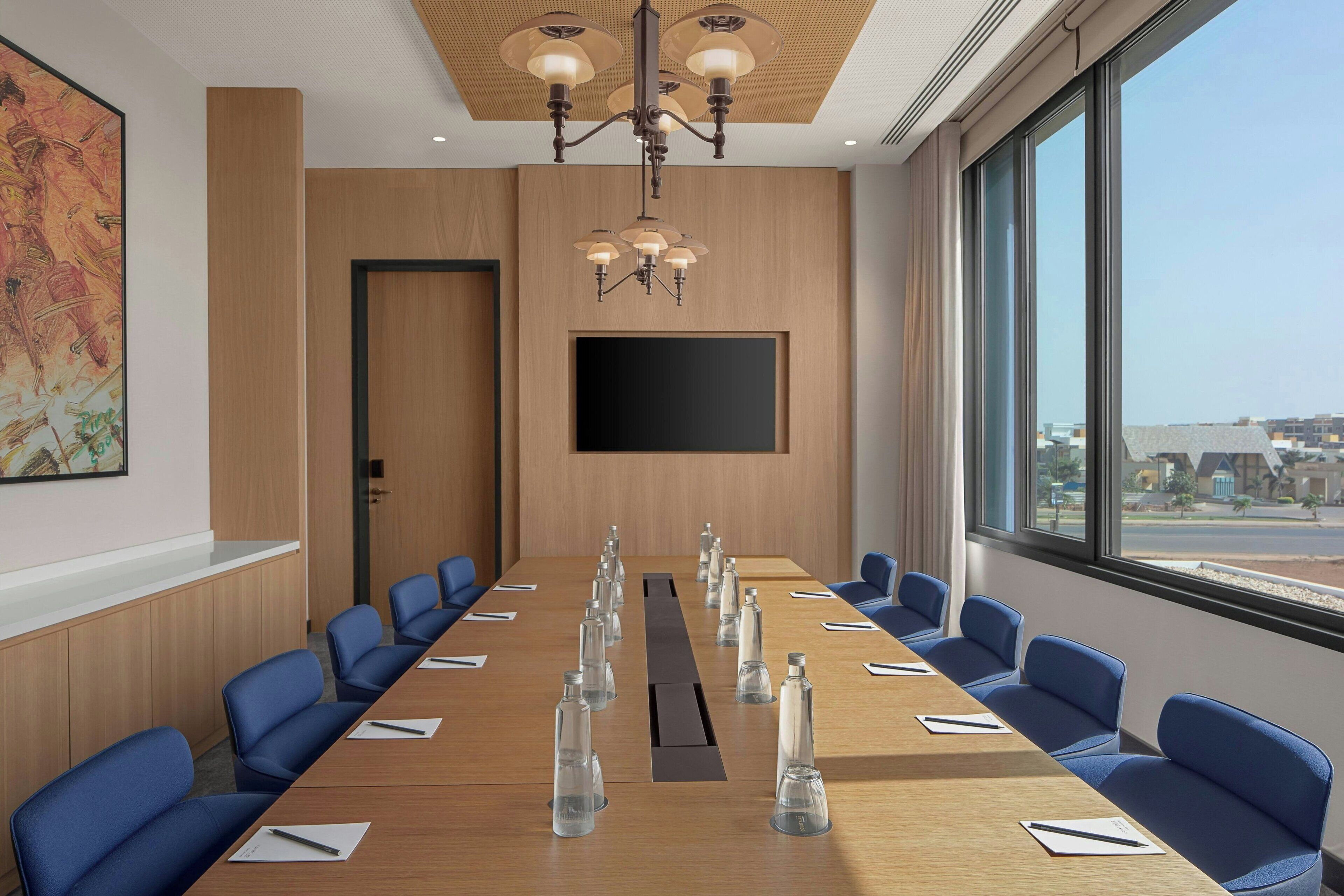 A conference room with a long table, blue chairs, a chandelier, a flat-screen TV, and large windows.