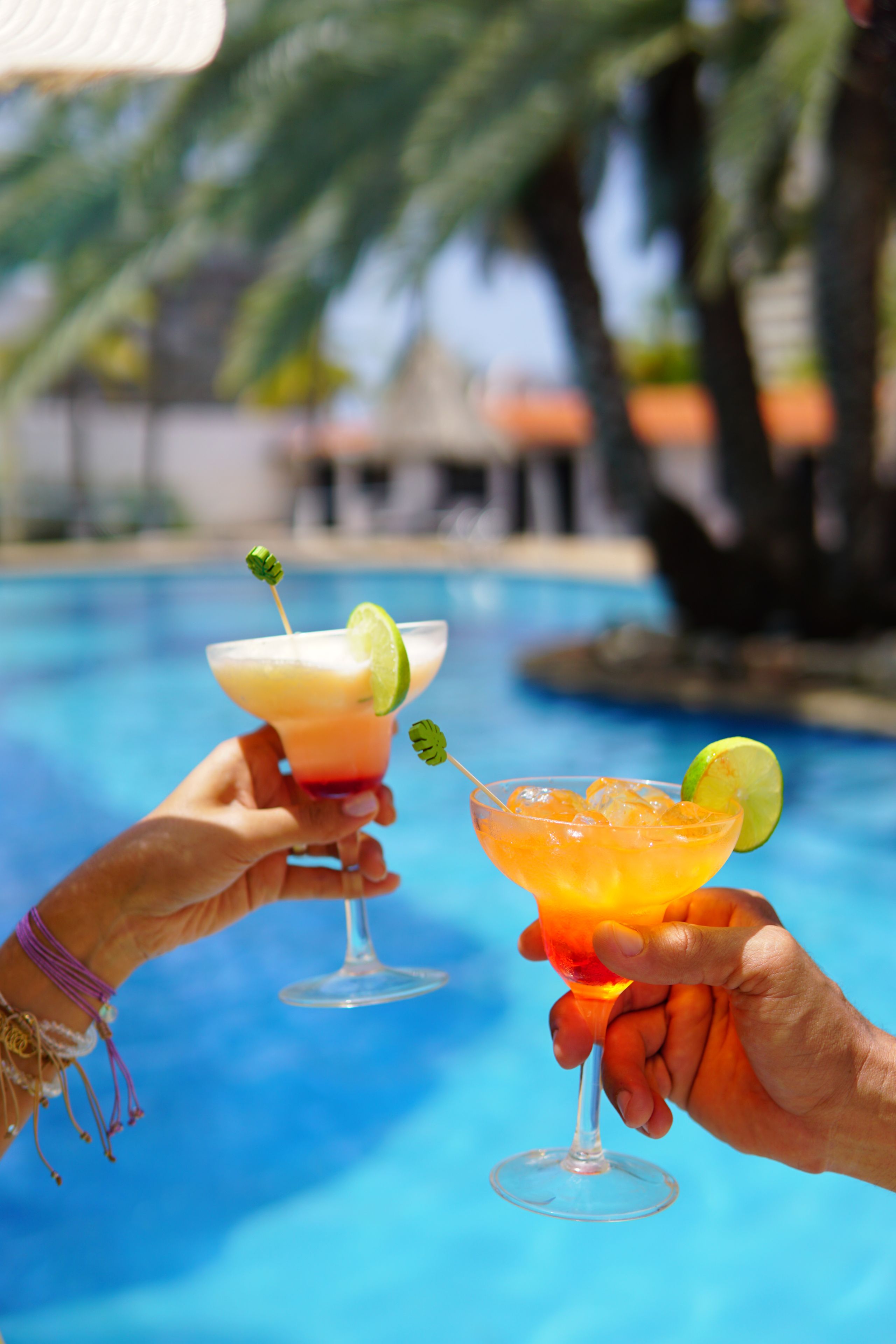 Zwei Hände halten Cocktailgläser mit Limettenscheiben, im Hintergrund ein Pool und Palmen.