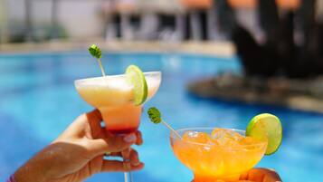 Zwei Hände halten Cocktailgläser mit Limettenscheiben, im Hintergrund ein Pool und Palmen.