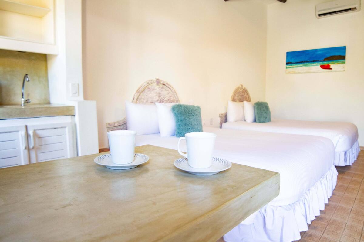 A hotel room with a wooden table, two beds, a sink, and a painting on the wall.