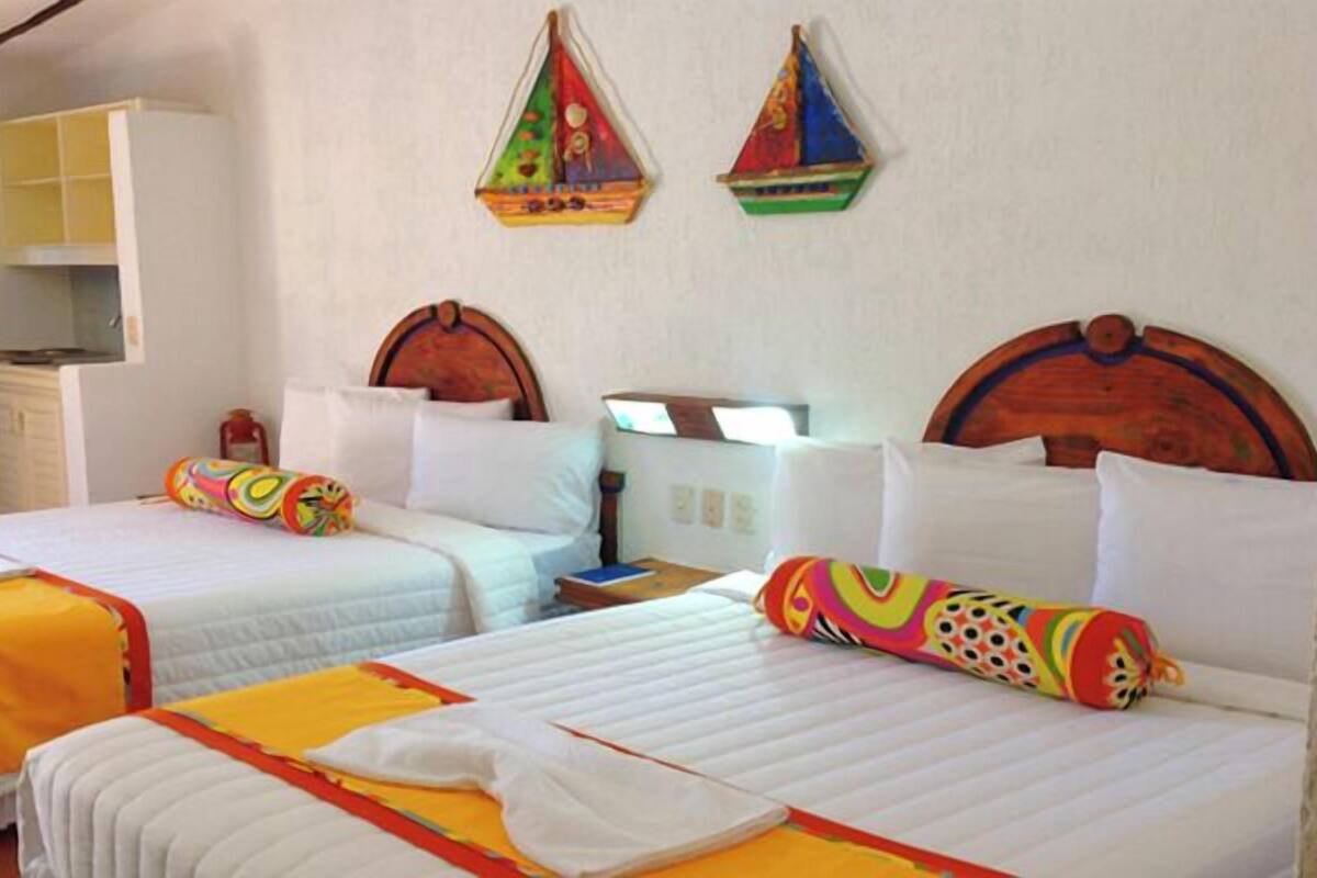 A hotel room with two beds, each with a colorful pillow and a white duvet. There are two wall-mounted decorations, a small desk, and a cabinet.