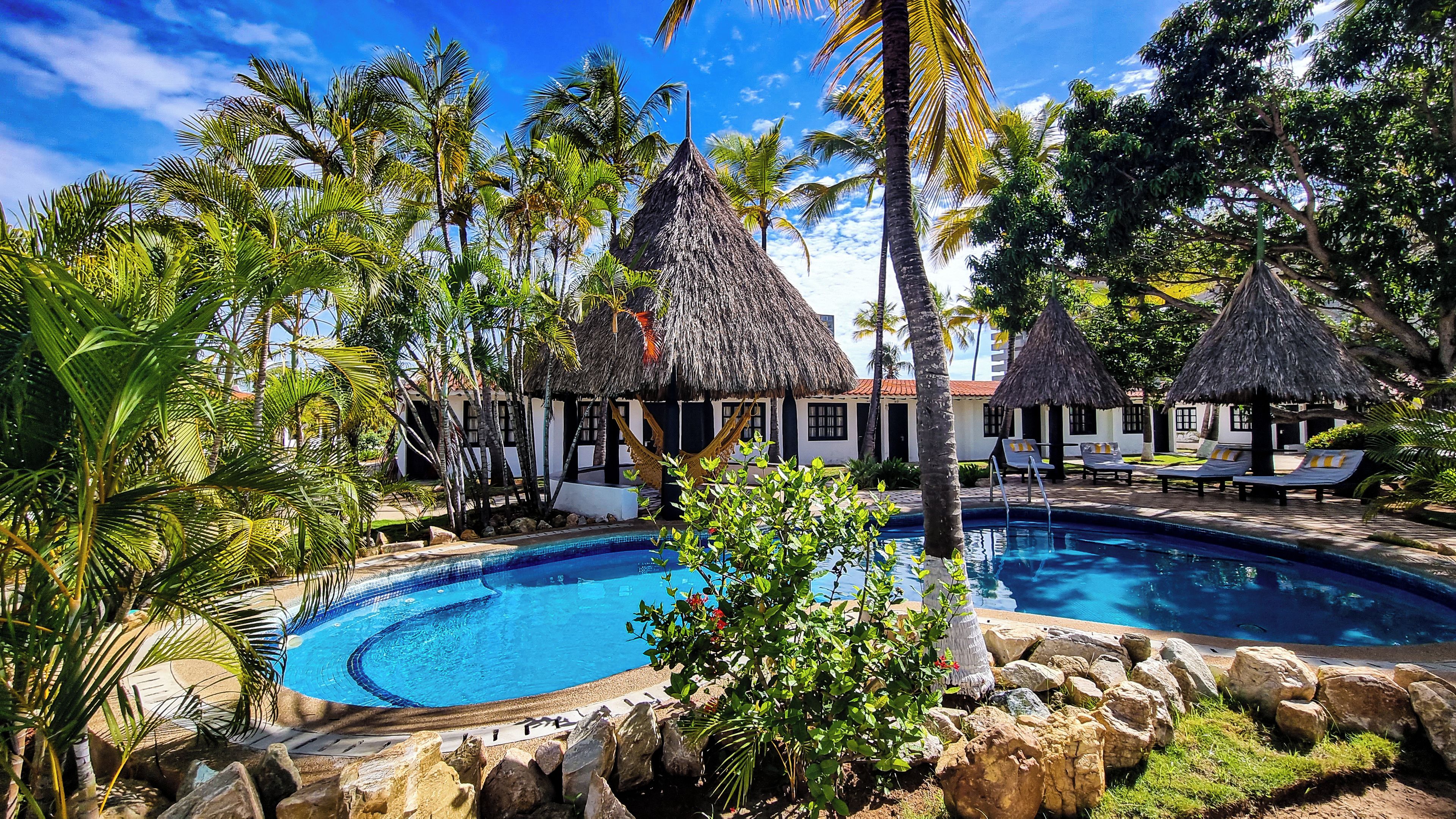 A resort with a swimming pool surrounded by palm trees and thatched-roof structures.