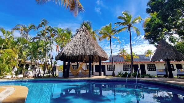 Una piscina con una estructura de techo de paja, palmeras y un edificio con techo de tejas.