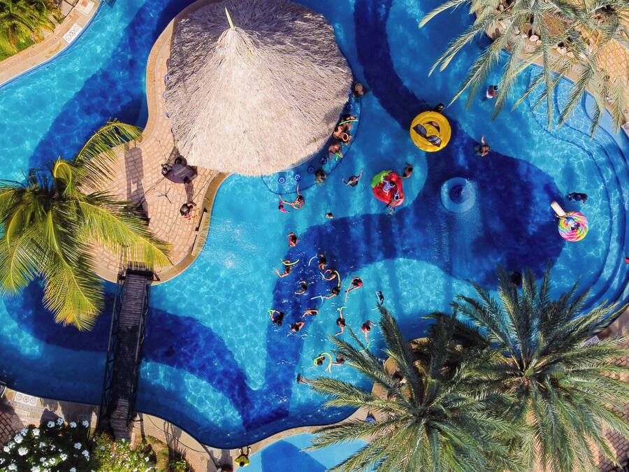An aerial view of a swimming pool area with multiple lanes, palm trees, and a thatched roof structure.