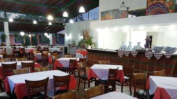 Un restaurant avec de nombreuses tables et chaises, un comptoir où sont empilées des assiettes et une fresque murale.