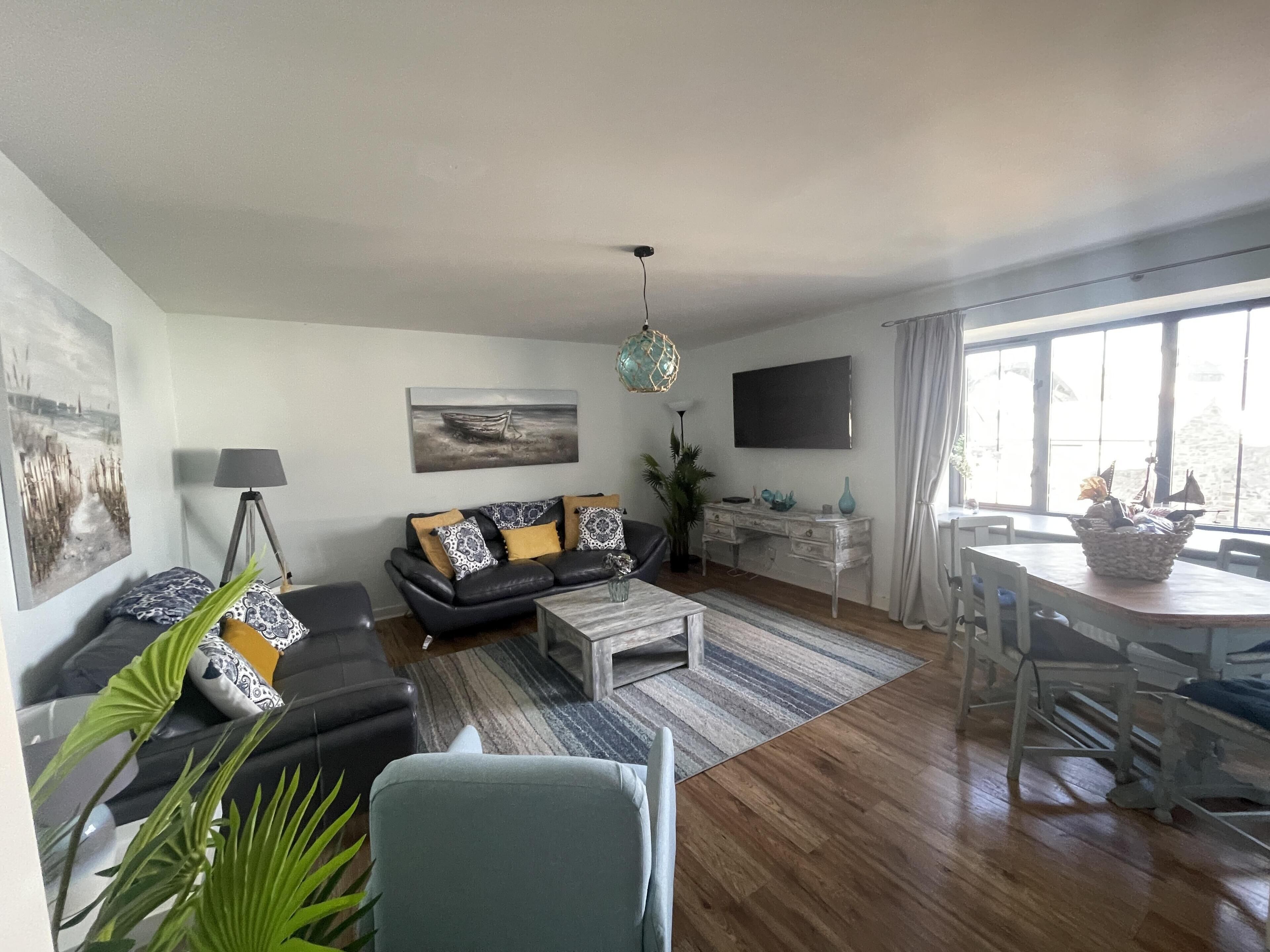 A living room with a sofa, coffee table, and dining table. There is a painting on the wall and a plant in the corner.