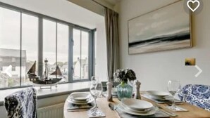 A dining area with a table set for two, a view of the city through large windows, and a painting on the wall. - Stunning Coastal Retreat -mins From Harbour/beach (St Austell)