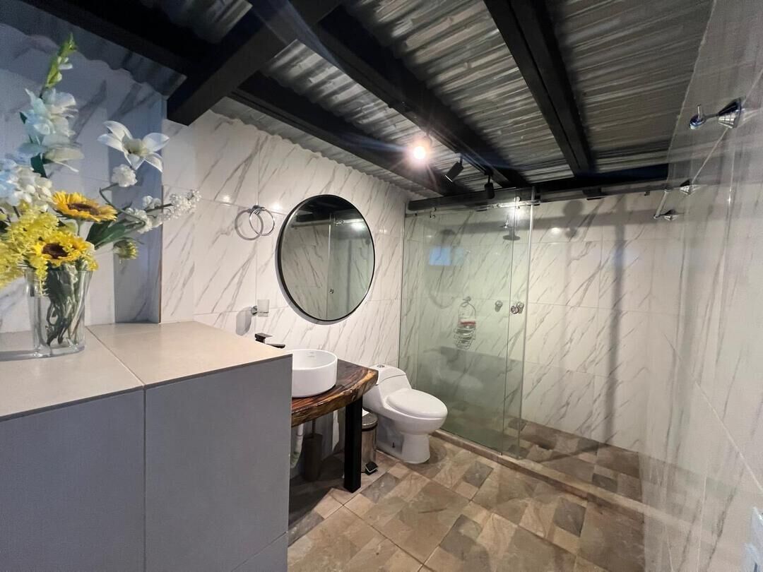 Un baño moderno con pared de mármol, un espejo redondo, un lavabo con encimera de madera, un inodoro y una ducha con mampara de vidrio.