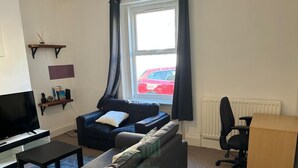 A living room with a sofa, a TV, a coffee table, a bookshelf, a window with curtains, and a radiator. - Cozy Coastal Getaway in North Shields (North Shields)