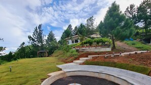 Um jardim com paisagismo, um gazebo, uma casa com telhado inclinado e uma estrutura de concreto curva.