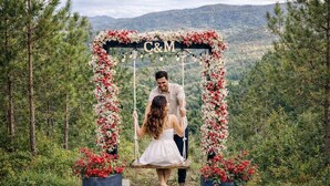 Um casal se balança sob um arco floral com as iniciais 'C&M', cercado por vegetação e montanhas.