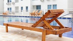 En solstol i trä placerad vid poolkanten, med en vit byggnad i bakgrunden.