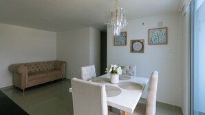 En matplats med ett vitt bord, beige stolar och en kristallkrona. I bakgrunden finns en beige soffa och på väggen hänger två inramade tavlor.