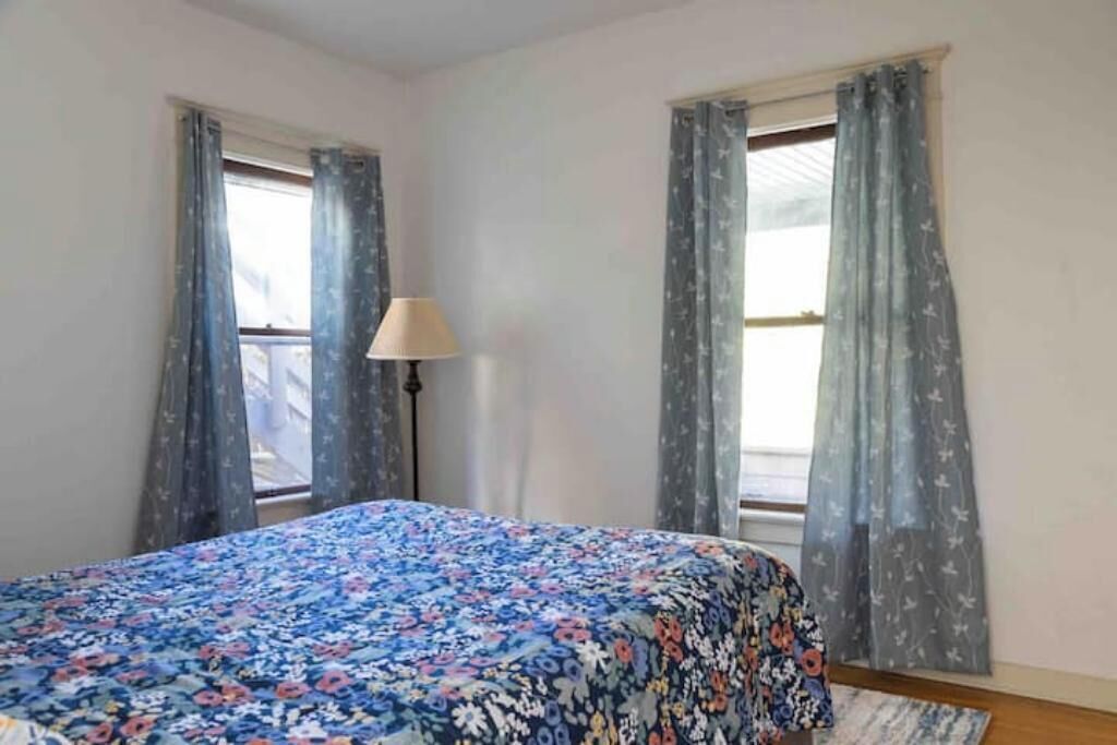A room with a bed, two windows with curtains, a lamp, and a rug.