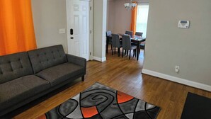 Una sala de estar con un sofá gris, un tapete estampado y una zona de comedor con una araña y sillas.