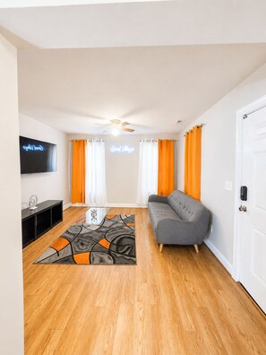 Uma sala de estar moderna com um sofá cinzento, piso de madeira, ventilador de teto e uma TV de tela plana fixada na parede.