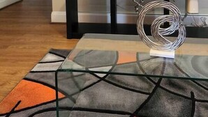 Un salón con un televisor de pantalla plana montado en la pared, una mesa de centro de cristal y un tapete geométrico, y un banco con una escultura de metal decorativa.