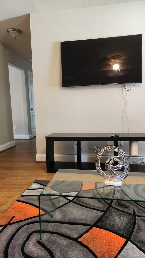 Uma sala de estar com televisão de tela plana na parede, mesa de centro de vidro e um tapete geométrico, além de um banco com uma escultura metálica decorativa.