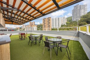 An outdoor terrace with artificial grass, metal tables, and chairs, overlooking a cityscape with high-rise buildings.