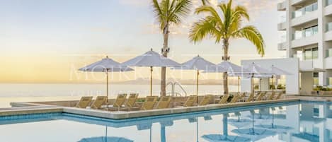 Zona junto a la piscina con sillones de descanso y sombrillas, palmeras y vista al océano.