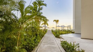 A pathway lined with palm trees leading to a building with a view of the ocean. - Luxury 2BR Beachfront Escape | Stunning Views from Private Balconies (Puerto Vallarta)