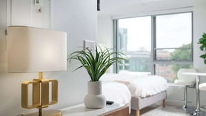 A modern hotel room with a bed, a bedside table with a lamp, a potted plant, and a view of the city through large windows. - Luxury Studio with King bed Gym near MIT Harvard in Central Square! (Cambridge)