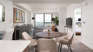 A modern living room with a gray sofa, a round wooden coffee table, and two beige armchairs. A large window offers a view of the cityscape. - Upscale 1BR MIT Harvard WiFi Gym W D Parking! (Cambridge)