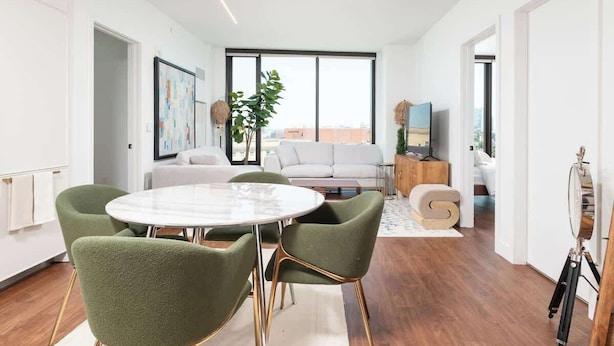 A modern living room with a round dining table, green upholstered chairs, a white sofa, and a large window offering a city view.