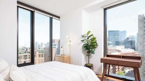 A bedroom with a large bed, a desk, a plant, and a view of the city through large windows.