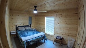 Een houten slaapkamer met een bed, een plafondventilator, een raam en een klein krukje.