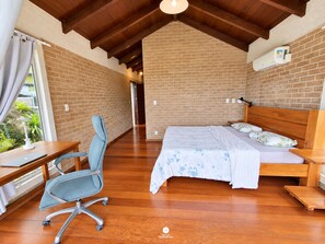A bedroom with a wooden ceiling, a brick wall, a bed with a floral bedspread, a desk with a chair, and an air conditioning unit. - Casa Garopaba Vista Panorâmica do Mar (Garopaba)