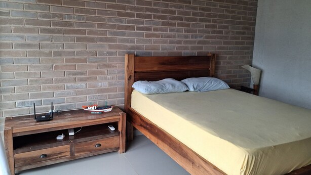 Una cama individual con cabecera de madera, una mesita de noche con lámpara y pared de ladrillo.