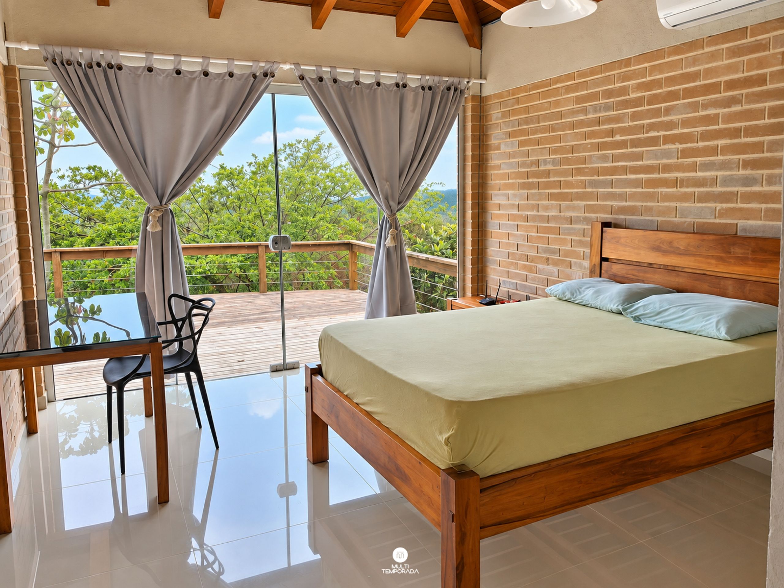 A bedroom with a wooden bed, a desk, and a chair, a balcony with a view of greenery, and a brick wall.