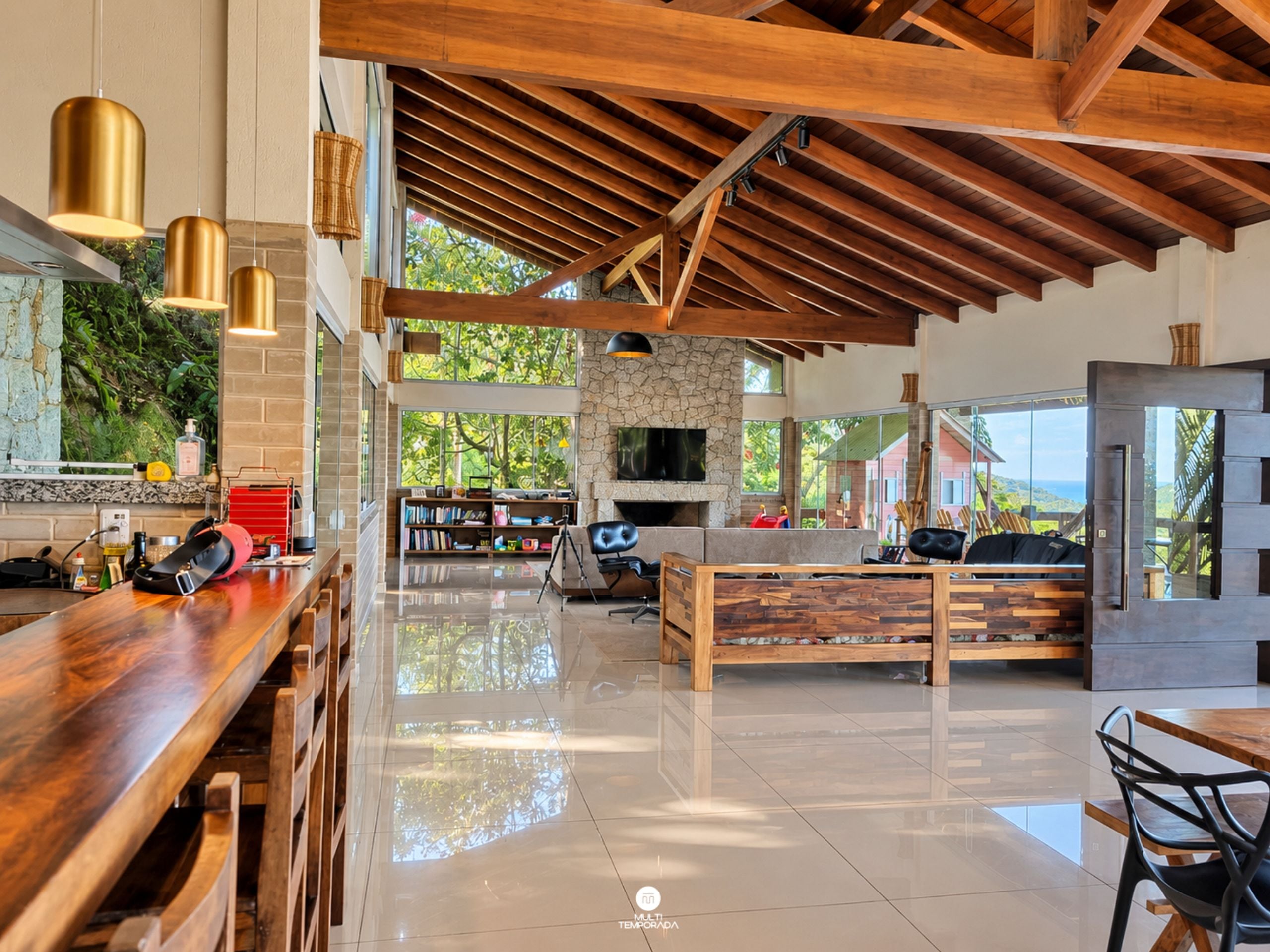 Amplia sala de estar con techo alto, vigas de madera, chimenea de piedra y un ventanal con vistas a la vegetación.