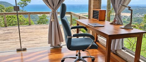A modern office space with a wooden desk, a chair, and a laptop. There is a large window with curtains, offering a view of the ocean and greenery.
