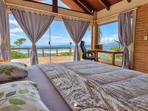 A bedroom with a bed, a desk, and a view of the beach. - Casa Garopaba Vista Panorâmica do Mar (Garopaba)