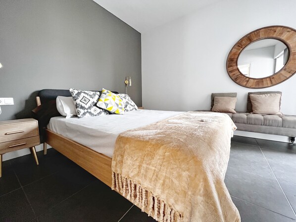 A modern bedroom with a large bed, a bedside table, a sofa, and a round mirror on the wall.