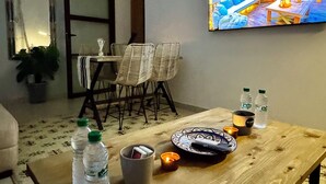 Un salon moderne avec une table basse en bois, deux chaises blanches, une plante en pot, une horloge et un grand écran de télévision qui diffuse un paysage pittoresque.