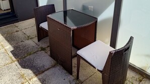 Een terras buiten met een glazen scherm, een tafel en twee rieten stoelen.