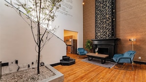 Un vestíbulo moderno con piso de madera, una chimenea negra, dos sillas azules, un pequeño árbol y una pared con patrones geométricos.
