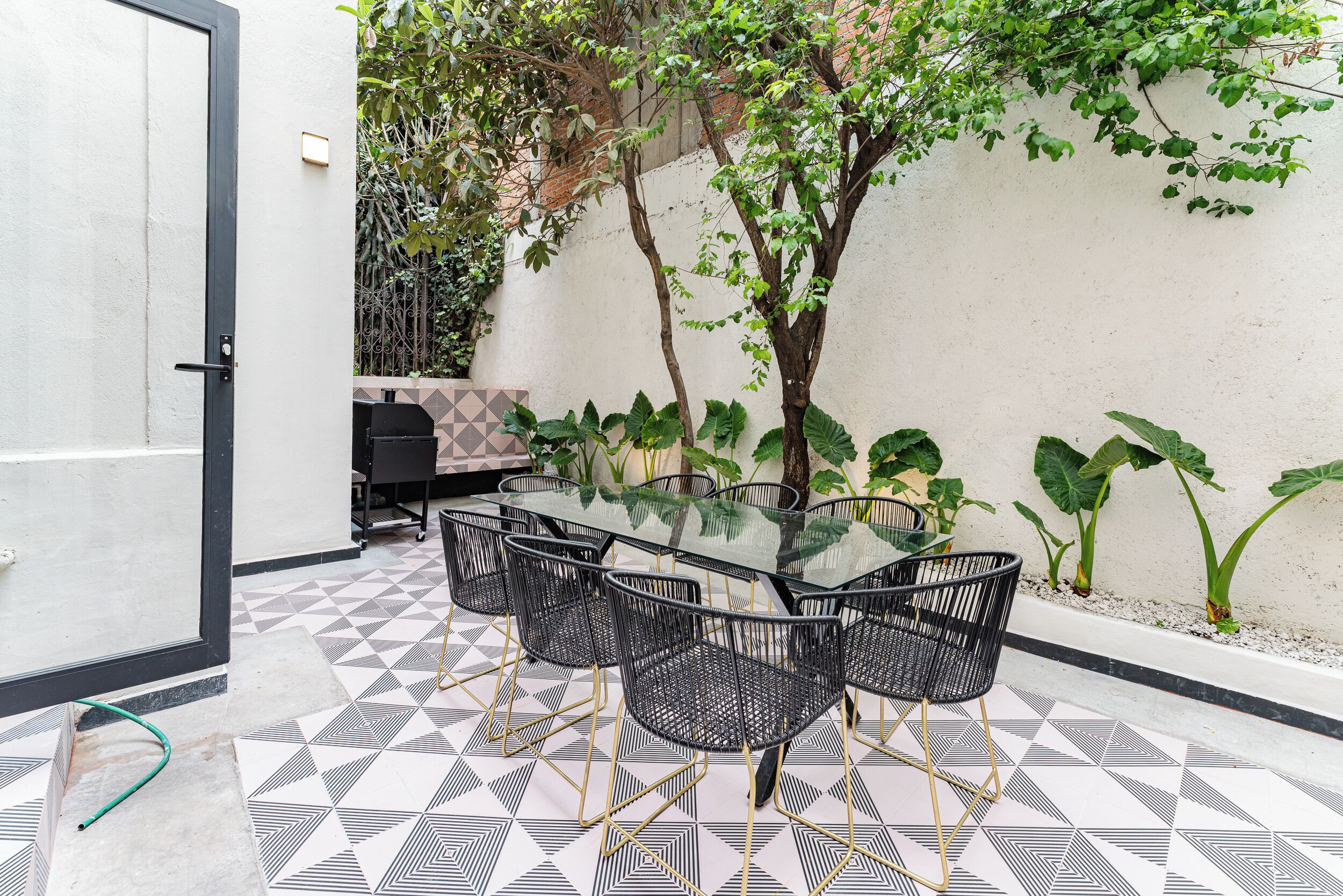 A patio with a glass-top table surrounded by black chairs, a tiled floor, and a white wall with greenery.